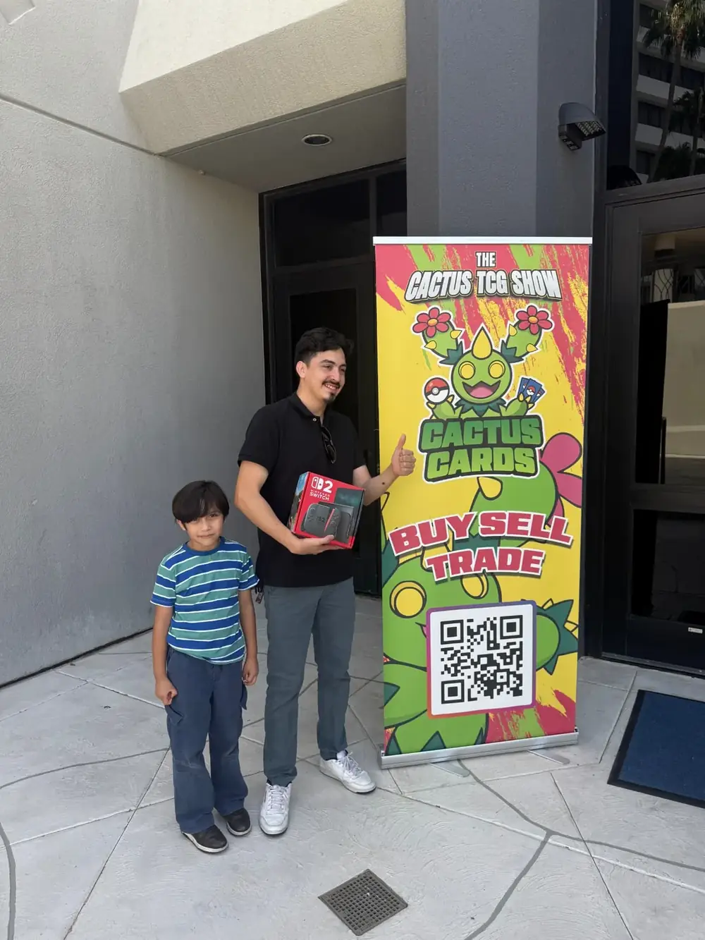 Nintendo Switch 2 giveaway winner and his son beside the Cactus TCG Show banner at the Mesa, Arizona card show