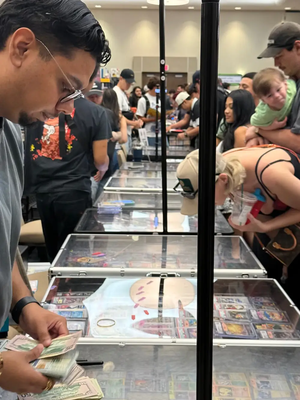 Vendor counting cash across glass cases of graded Pokémon cards while families browse The Cactus TCG Show in Mesa, Arizona